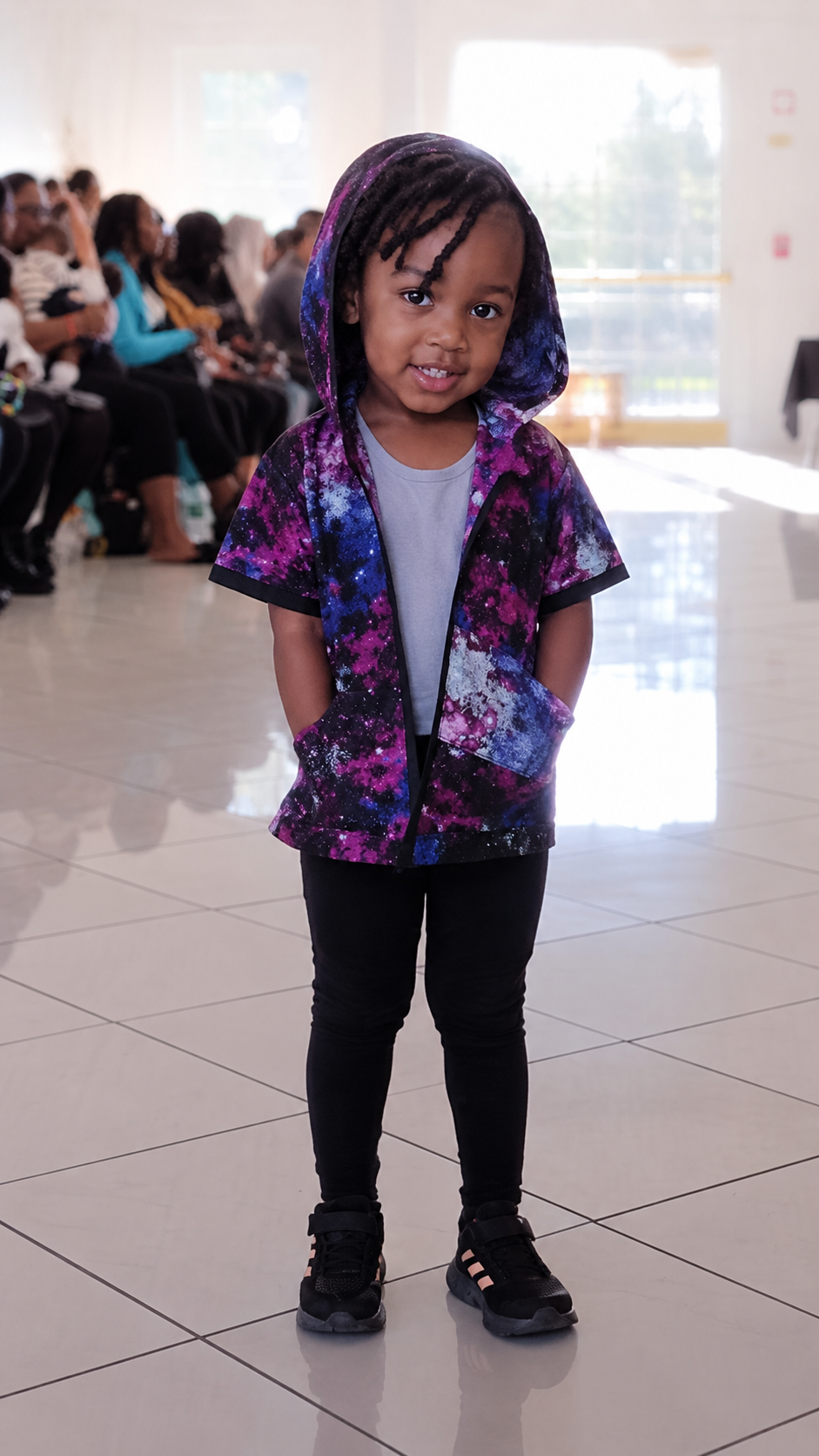 A young child walking down a tiled runway wearing a short-sleeve hooded shirt with a purple, pink, and blue galaxy print, black pants, and black sneakers.