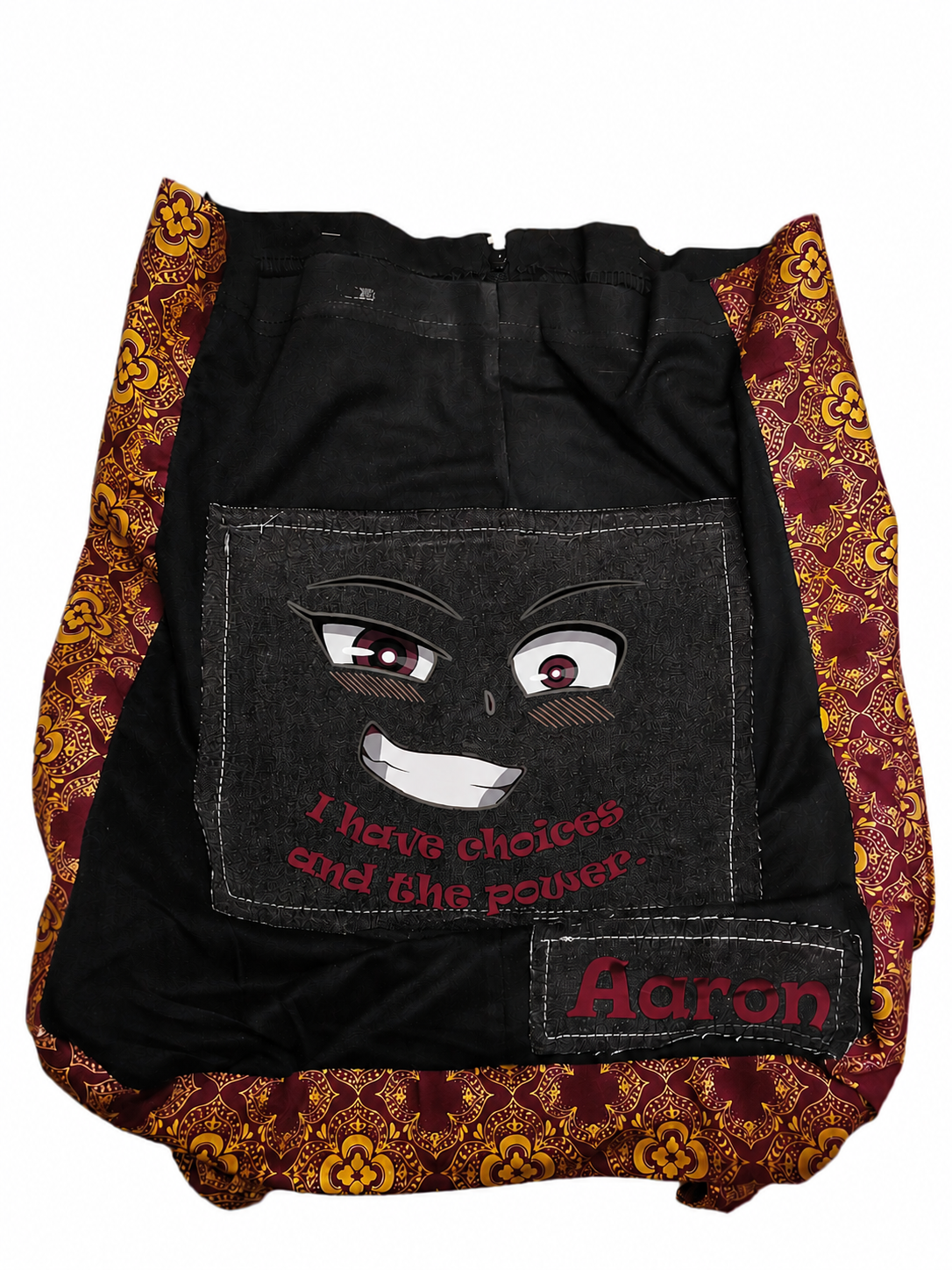 A black denim backpack featuring a graphic of a smiling face with red eyes and the text 'I have choices and the power' on the front pocket, with a 'Aaron' name patch on the lower right. The backpack has patterned burgundy and gold fabric trim on the sides and bottom.
