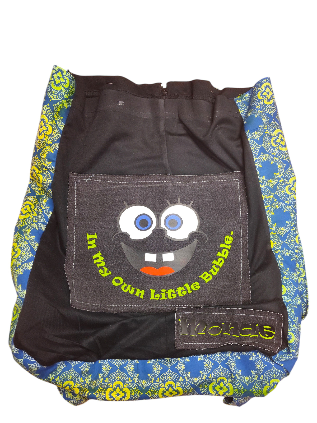 A handmade fabric backpack or bag featuring a black front pocket with a cartoon face patch and the text 'In My Own Little Bubble', surrounded by blue and yellow patterned fabric sides.