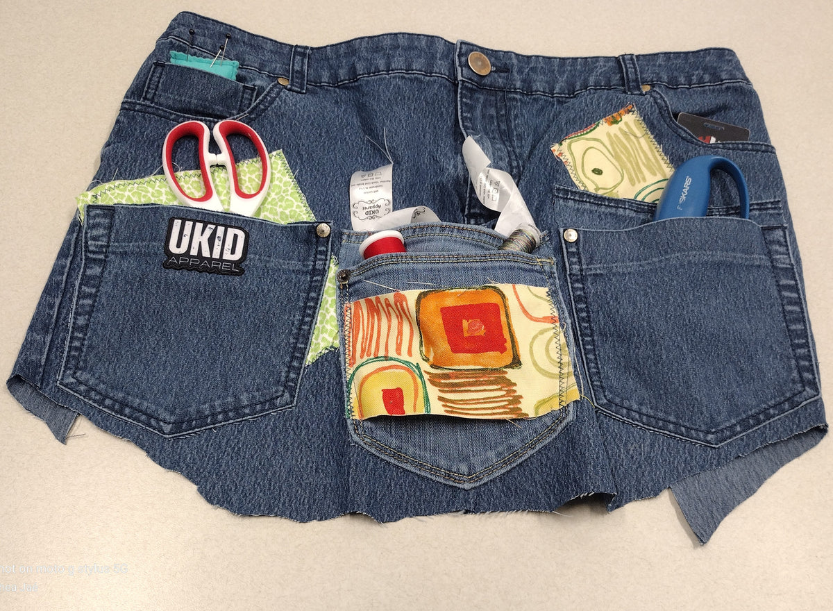 A pair of blue denim shorts with frayed hems, featuring colorful patchwork fabric inserts in the back pockets and various tools like scissors and a utility knife tucked into the pockets. A 'UKID' brand patch is visible on the left pocket.