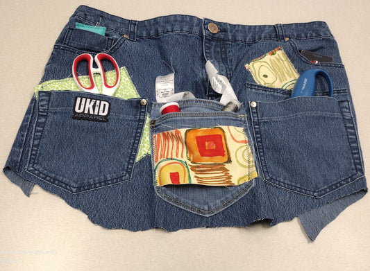 A pair of blue denim shorts with frayed hems, featuring colorful patchwork fabric inserts in the back pockets and various tools like scissors and a utility knife tucked into the pockets. A 'UKID' brand patch is visible on the left pocket.