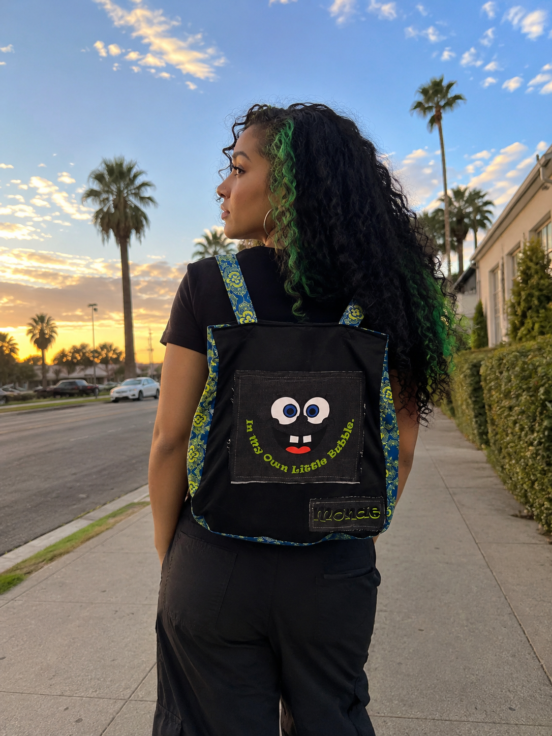 A woman standing on a sidewalk outdoors at sunset, wearing a black backpack with a cartoon face patch and blue-green patterned trim. Palm trees, a street, and a building are visible in the background.