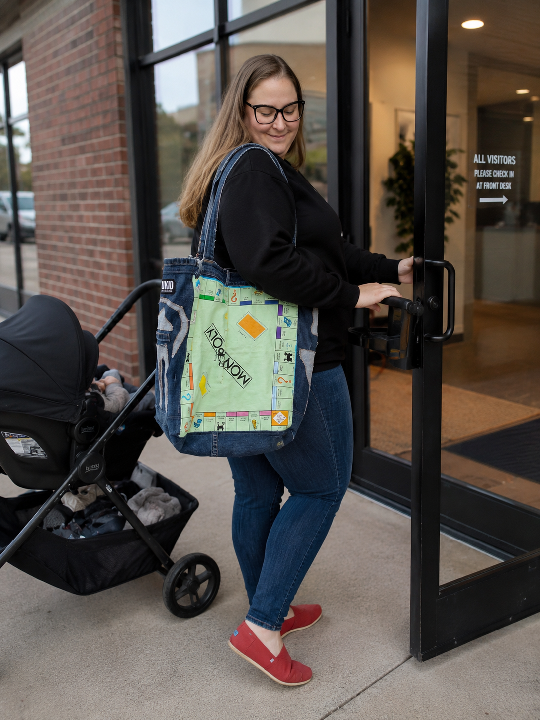 A woman wearing glasses, a black long-sleeve shirt, and blue jeans carries a denim tote bag with a colorful Monopoly game board design on the front. She is standing outside a building next to a black stroller, holding the door handle.