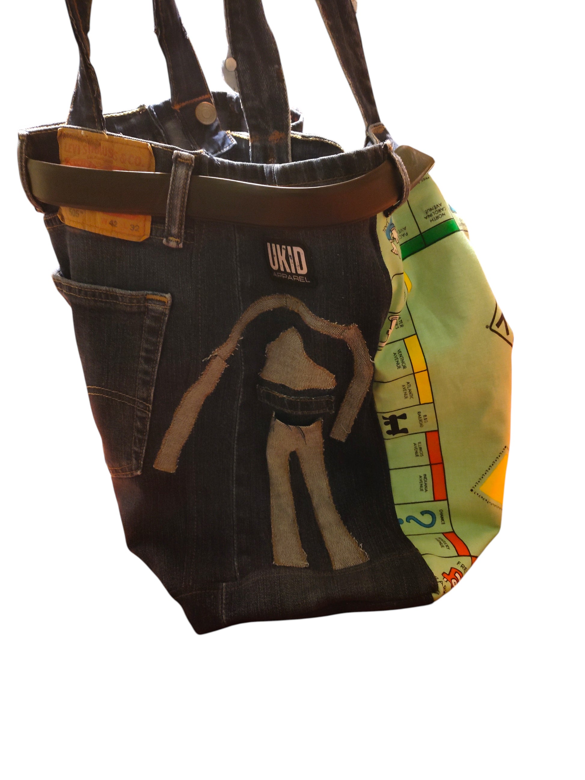 A tote bag made from repurposed denim jeans, featuring a dark blue denim body with a beige graphic of a person, a brown leather-like waist strap, and a green Monopoly game-board style side panel.