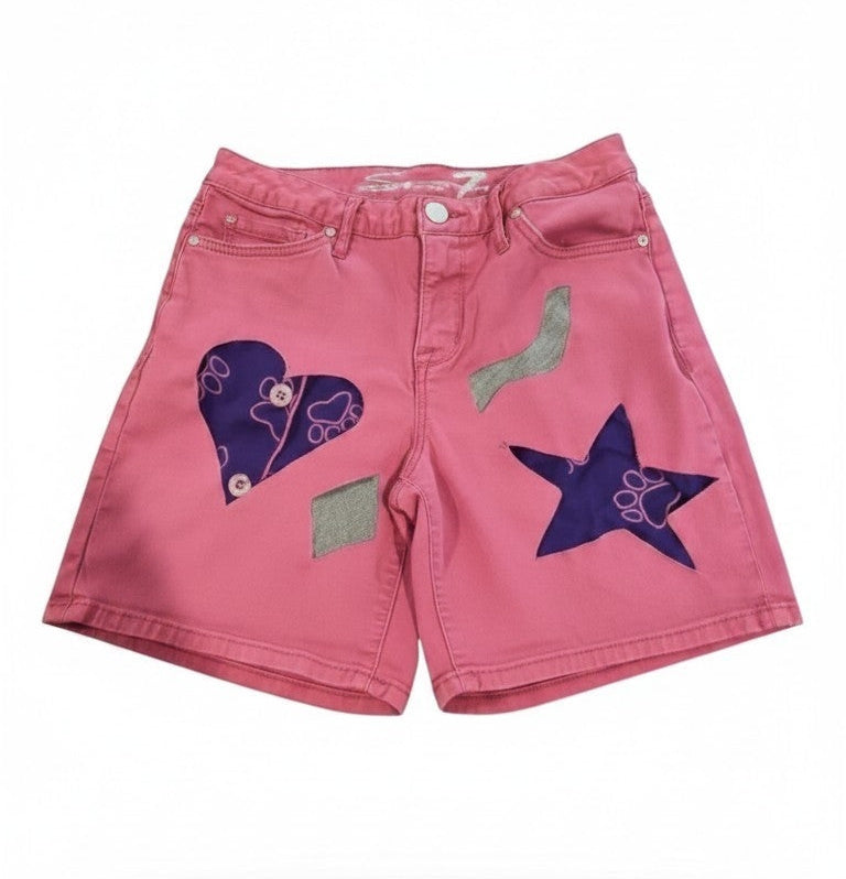 Pink denim shorts with decorative applique patches, including a purple heart, a purple star, and a green leaf, shown against a plain white background.